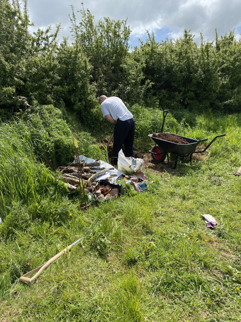 Man building slow worm hibernaculum at Cobthorn reserve. Digging near bush with logs and wheelbarrow nearby.